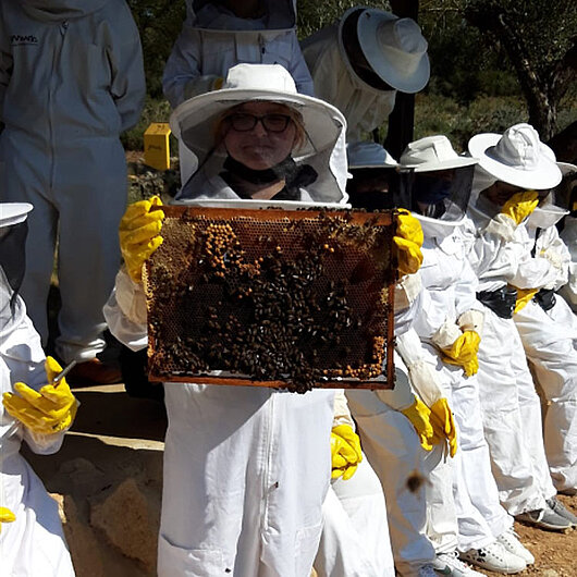 Jugendliche in Imkerschutzkleidung, eine hält eine Bienenwabe in die Kamera