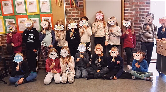 Eine gruppe von Grundschülerinnen und Grundschülern. Die Kinder halten sich Masken in Form von unterschiedlichen Tierköpfen vor das Gesicht.