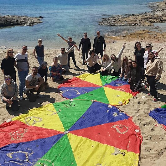 Gruppe mit 20 Erwachsenen an einem Strand steht rund um zwei bunte Fallschirme, die im Sand liegen.