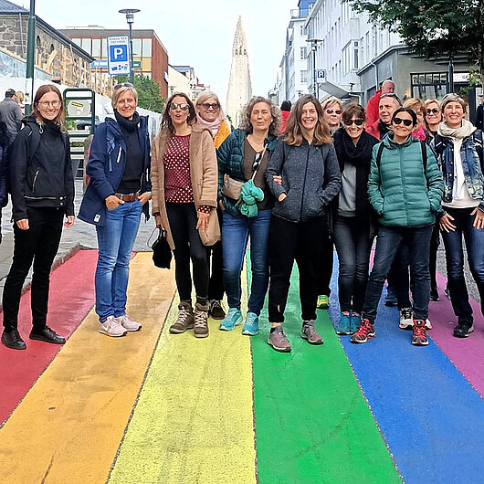 Gruppe von Frauen stehen nebeneinander in städtischer Umgebung und blicken lachend in die Kamera. Sie stehen auf farbigen Streifen in Regenbogenfarben. 