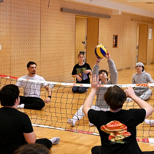 Schüler:nnen in der Sporthalle üben Volleyball im Sitzen