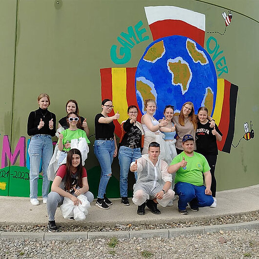 Gruppe Jugendlicher steht vor einem Wandgemälde. Man sieht eine bunte Weltkugel und die Flaggen der Schulpartnerschaft: Spanien, Deutschland und Tschechien. An der Seite ist eine Biene gemalt.