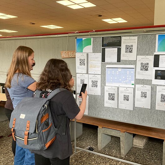 Schülerinnen und Schüler scannen mit Smartphones QR-Codes auf einer Stellwand